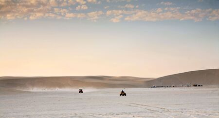 Quad-bikers play at sunset on the salt-flats beside the sand dunes at Mesaieed, Qatar. (some motion blur on the nearest biker). A tent encampment stands at the foot of the dunes.の写真素材