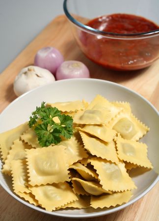 Fresh ravioli, ready for cooking, with a bowl of tomato sauce, onion and garlic on a wooden board.の写真素材