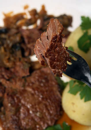 A chunk of braised steak on a fork, close-up, with the rest of the meal out of focus in the...