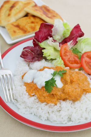 Chicken tikka masala topped with fresh yoghurt on a bed of basmati rice, all served with a fresh salad, with naan bread in the backgroundの写真素材
