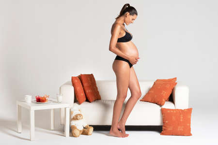 Young beautiful pregnant woman standing close to a white sofa holding hands on her belly dressing underwearの写真素材
