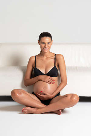 Young beautiful pregnant woman sitting close to a white sofa holding hands on her belly dressing underwearの写真素材