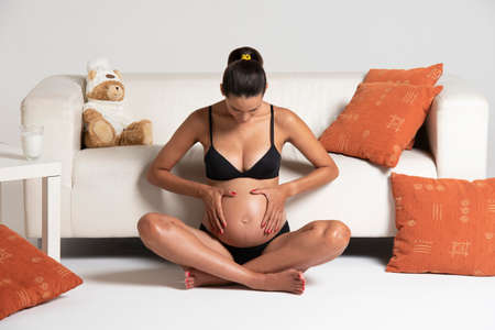 Young beautiful pregnant woman sitting close to a white sofa holding hands on her belly dressing underwearの写真素材