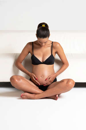 Young beautiful pregnant woman sitting close to a white sofa holding hands on her belly dressing underwearの写真素材