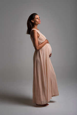 Young beautiful pregnant woman standing on the floor wearing a long pink dress vertical view grey backgroundの写真素材