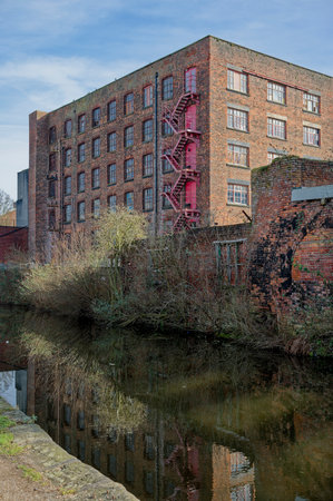 Historic brick factory mill building with red fire escape beside a calm canal.の写真素材