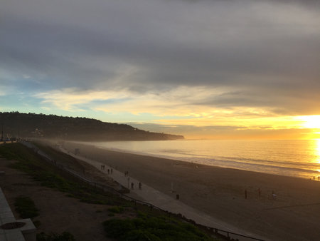 Sunset Casts a Warm Glow Over the Beach as People Stroll Along the Shoreline Near a Coastal Pathの写真素材