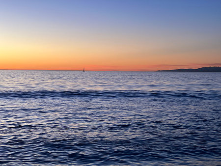 Vibrant Sunset Over the Calm Ocean With a Distant Sailboat on the Horizonの写真素材