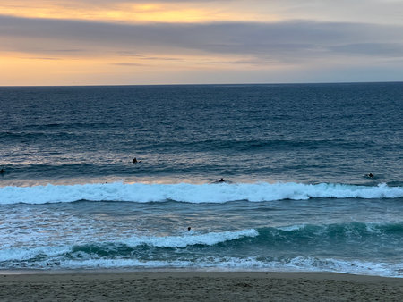 Surfers Enjoying the Evening Waves at a Peaceful Beach Under a Vibrant Sunset Skyの写真素材