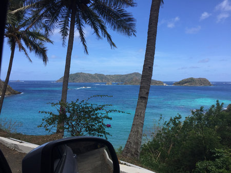 View of Serene Turquoise Waters and Distant Islands Framed by Palm Trees Along a Coastal Roadの写真素材