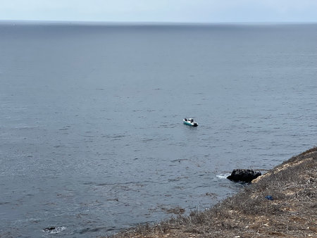 Fishing Boat Drifts Peacefully on Calm Waters Near Rocky Coastline Under a Cloudy Skyの写真素材