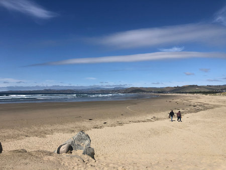 Two People Stroll Along the Sandy Beach Under a Clear Blue Sky With Rolling Waves in the Background Near a Coastal Townの写真素材