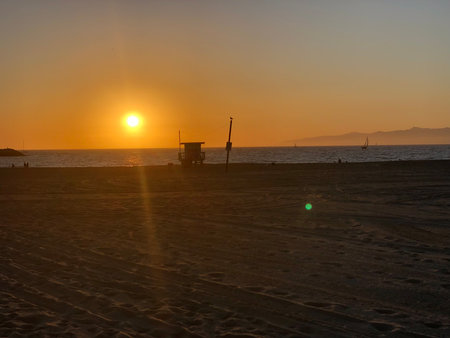 Sunset Over the Quiet Beach With Silhouettes of a Lifeguard Tower and Distant Sailboatsの写真素材