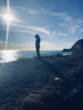 Person Standing on Rocky Shore Gazing at the Sunlit Ocean Under a Vast Blue Skyの写真素材