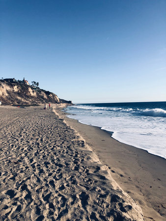 Waves Gently Lap at Sandy Shore While Sunlight Illuminates Coastal Cliffs in a Tranquil Afternoon Sceneの写真素材
