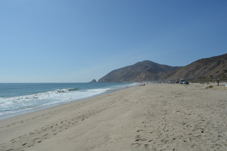 Serene Beach Landscape With Golden Sand, Gentle Waves, and Distant Mountains Under a Bright Blue Skyの写真素材