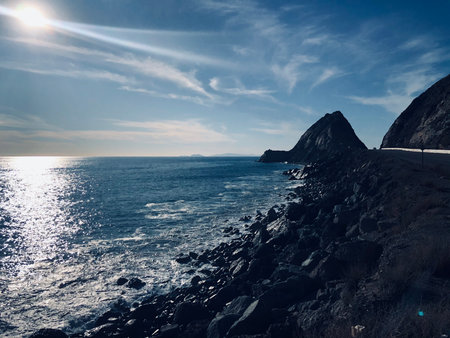 Sparkling Sea Waves Lap Against Rugged Coastal Cliffs Under a Bright Sun at Midday Near Serene Blue Watersの写真素材