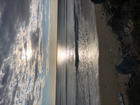 Gentle Waves Lap at the Sandy Shore as the Sun Sets Behind a Blanket of Cloudsの写真素材