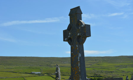 Celtic Cross Stands Tall Against a Backdrop of Green Hills Under a Clear Blue Sky in a Serene Rural Settingの写真素材