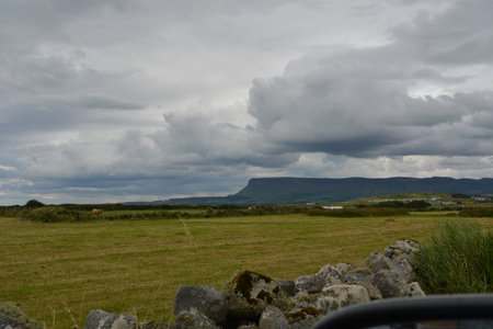 Vast Green Fields Stretch Beneath a Dramatic Sky as Rolling Clouds Gather Over Distant Hills in a Tranquil Rural Landscapeの写真素材