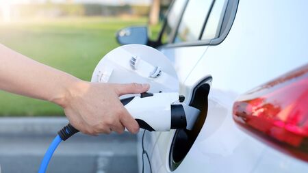 Electric car or ev is charging at station . man use the white power cable and plug on nature background. eco and clean energy concept with the mobile phone smart technology. zero gas and no transmission.の写真素材