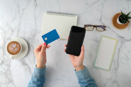 Flat lay of Woman hand with credit card and mobile phone with coffee cup, smartphone, computer, notebook, planner and stationary on marble table background. Top view. modern style. Digital online shopping and banking conceptの写真素材
