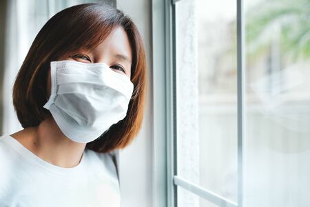 smiling woman stay home wear mask and looking at the window for self quarantine to stop the corona virus, covid19 pandemic outbreak . keep social distancing by call friend and doctor.の写真素材