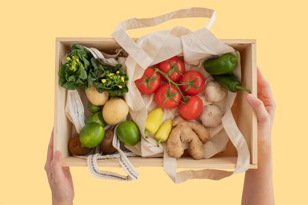 hand hold the pine wood box and cotton eco bag with fresh vegetables flat lay on yellow background.plastic free for groceries product shopping and delivery. zero waste lifestyle .の写真素材