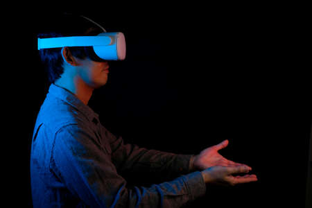 The Asian man is wearing VR headset. He is holding his hand to receive something in virtual reality. Blue neon theme and black background.の写真素材