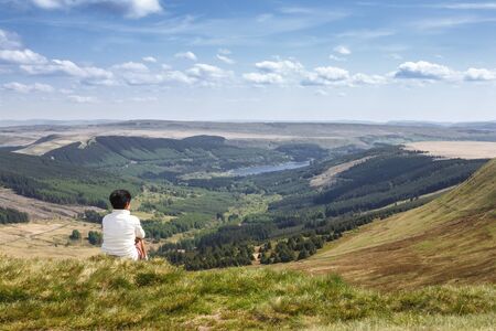 Indian woman sitting on a hill in Welsh countryside looking at a valley and toward the horizon. Pen Y Fan, Brecon Beacons, South Wales, UKの写真素材
