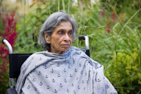 Old woman, British Asian Indian, sitting in a wheelchair looking sad and worried. UK mental health concept, depression, healthcare and caring for the elderlyの写真素材