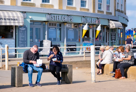 WHITBY, UK - September 21, 2022. People sitting eating fish and chips on a bench outside a fish and chip shop, Whitby Harbour, North Yorkshireのeditorial素材