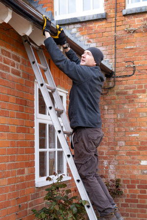 Man on a ladder fixing fitting a rain gutter on the eaves of a UK houseの写真素材