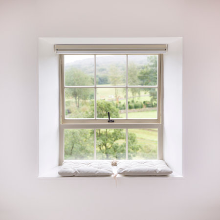 Traditional wooden sash window with a cushioned window seat, set in white walls in a UK home, with a scenic view of countrysideの写真素材