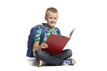 8 year old school boy with backpack sitting reading a red book on white backgroundの写真素材
