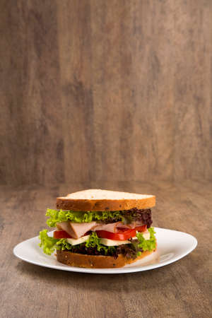 Sandwich on a white plate with turkey breast, tomato, lettuce and cheese on the tableの写真素材