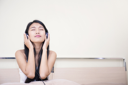 Asian woman listening to music with her headphones while sitting on her bedの写真素材
