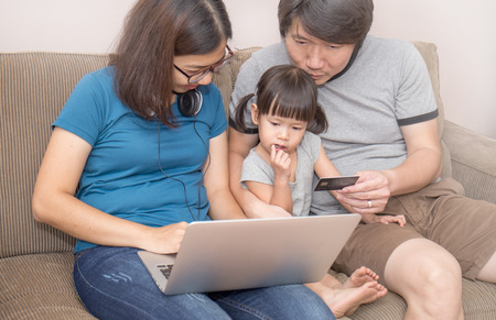 Asain parents and their daughter are doing shopping online using laptop and smiling, sitting on the sofa at home. Father is holding credit cardの写真素材