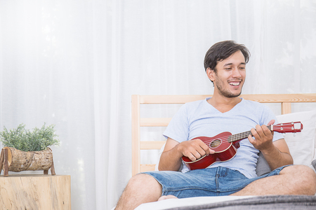 Young man sitting on bed, playing a ukuleleの写真素材