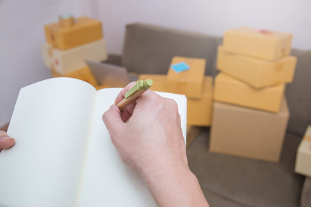 Close up hand holding pen writing note on a book. Start up small business entrepreneur SME working with box at home concept, online marketing packaging box and delivery, SME conceptの写真素材