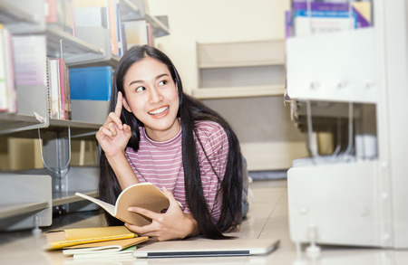 Attractive asian student(woman) lying in between bookshelves in modern university library reading a...