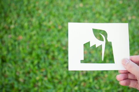 Hands holding cut paper with the  factory shape over green glass background. friendly green living. Sign and symbol background banner template of Earth day conceptの写真素材