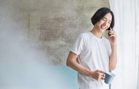 Portrait of young handsome asian man hand on smartphone and coffee mug in bedroom. Smile happy asian boy in bed morning lifestyle near window. Japanese man with technology conceptの写真素材