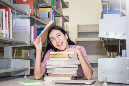 Attractive asian student ( woman ) sit between bookshelves in modern university library hands...