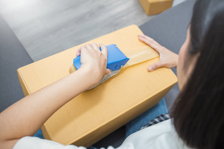 Young asian woman taping up a cardboard box in home office SME e-commerce business, relocation and...