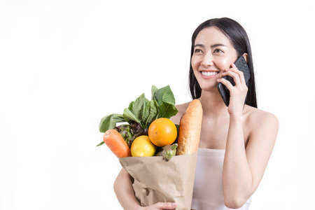 Closeup portrait of beautiful asian woman holding mobile phone grocery vegetables and food isolated on white background. Healthy lifestyle shopping online e-commerce telemarketing delivery concept.の写真素材