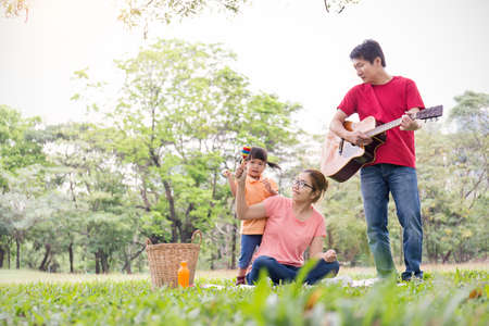 Portrait of happy family man woman and little girl playing guitar and maracas outside. Young family of three having fun picnic together outdoor. Asian parents and girl jumping happy and smile. Happiness and harmony in life.の写真素材