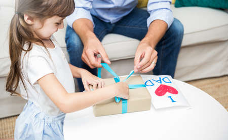 Little caucasian girl daughter congratulate daddy giving a surprise card and gift box. Happy family holiday father and girl celebrate together. Love lifestyle unwrap boxing or fatherâs day conceptの写真素材