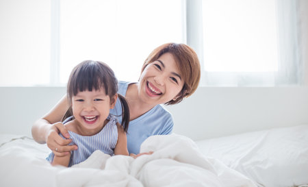 Portrait of happy asian mother and daughter laughing bonding in bed. Mommy and little girl spending time at home. Fun love family lifestyle together motherâs day conceptの写真素材