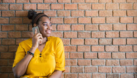 Close up portrait of happy confident african black woman talking on the mobile cell phone isolated on orange brick wall. Confident modern university student or business girl, technology communication connection conceptの写真素材